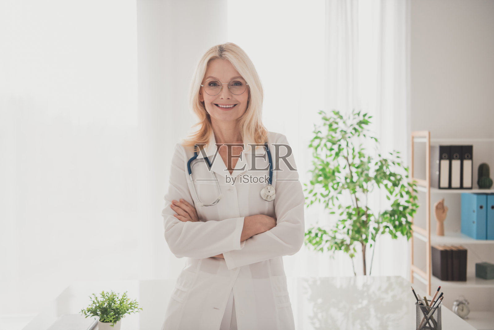 自信的女性高级医生站在医院诊所办公室，穿着白色制服，佩戴听诊器，双臂交叉。照片摄影图片