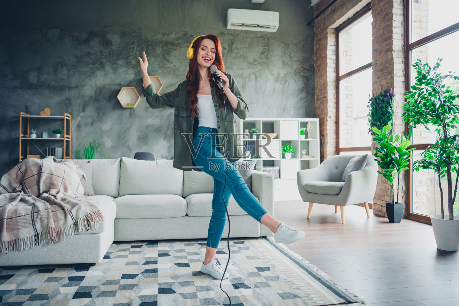 全身照：一位迷人的年轻红发女性在家居舒适的客厅里穿着卡其色服装，手持麦克风唱卡拉OK。照片摄影图片