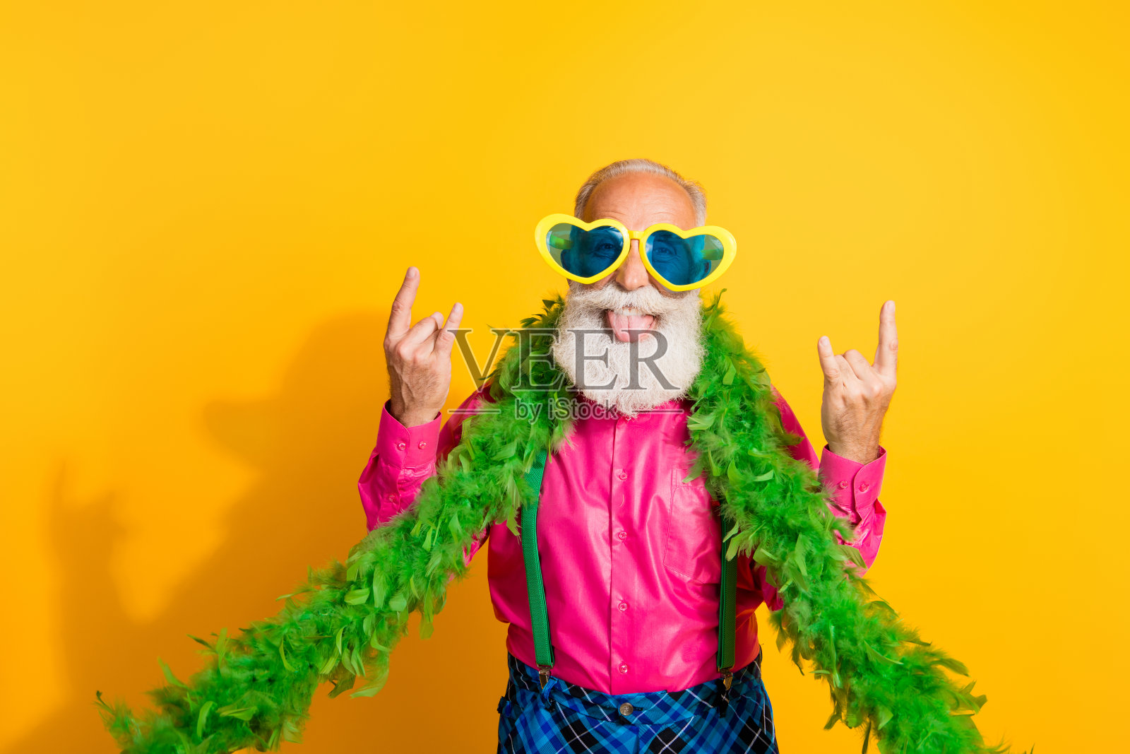 穿着粉色衬衫和复古格子裤的帅气退休男士，做出摇滚手势，身穿华丽服装，背景为黄色。照片摄影图片