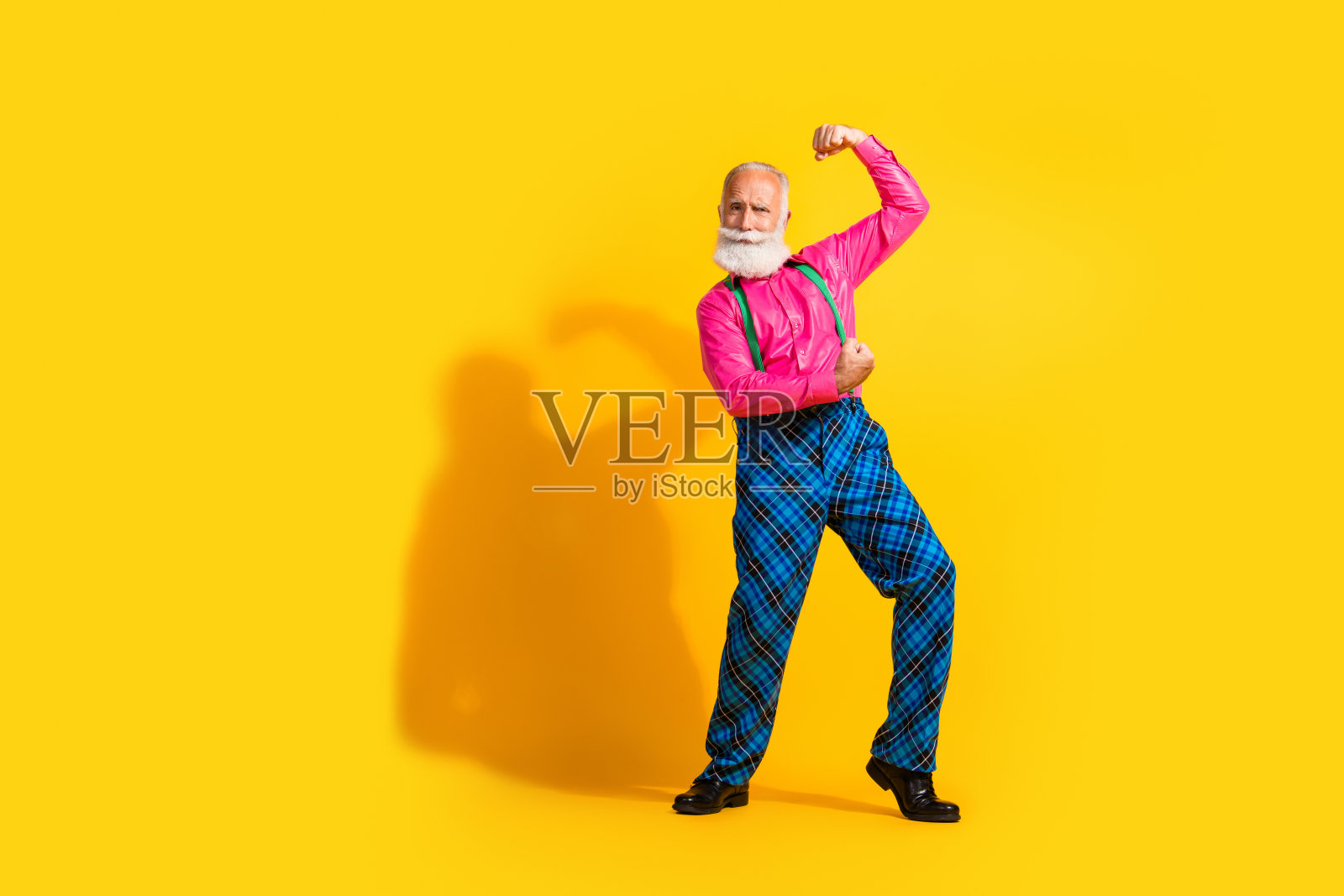 全身照，留着胡子的绅士在充满活力的黄色背景下展示肌肉，穿着时尚的吊带裤。照片摄影图片