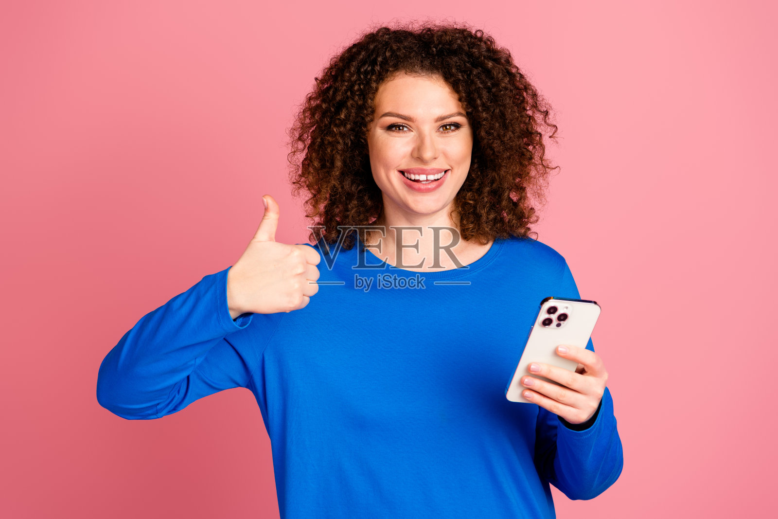 一位快乐的年轻女性，卷发，背景为粉色，举起大拇指，手中拿着智能手机，表达积极和成功的情绪。照片摄影图片