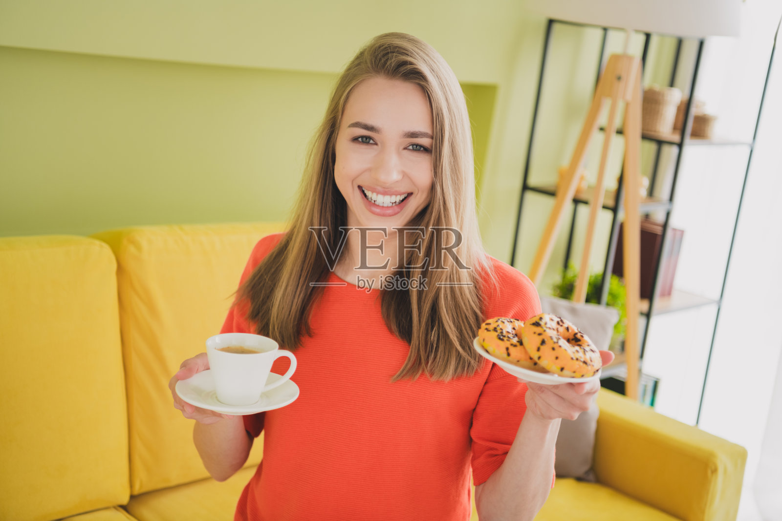 可爱的年轻女孩在宽敞的室内房间里穿着橙色衣服，手里拿着咖啡和甜甜圈，春天的气息弥漫。照片摄影图片
