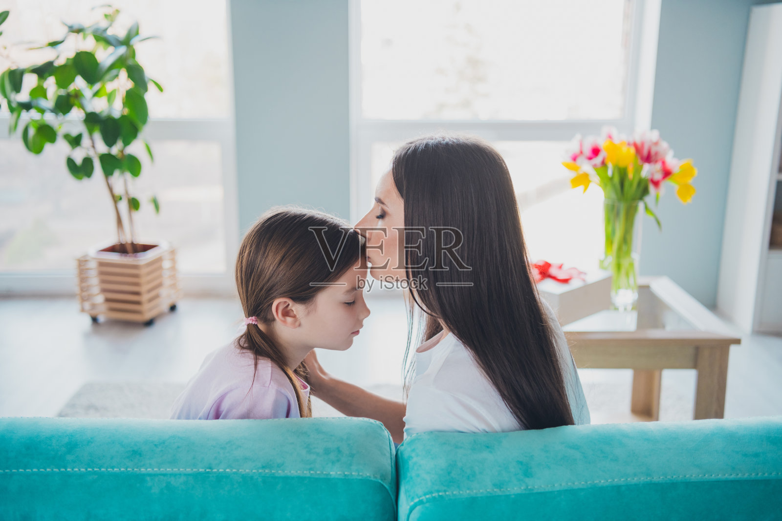 温柔可爱的妈妈在周末时光中亲吻她的女儿，坐在家里的沙发上。照片摄影图片