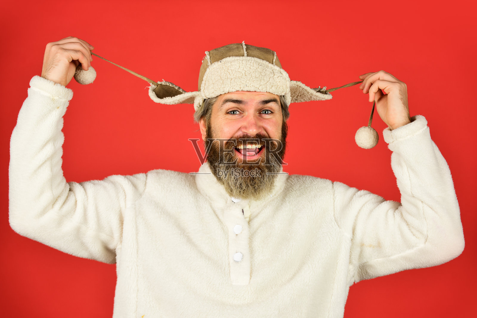 有胡子的搞笑嬉皮士。冬季时尚概念。适合寒冷天气的保暖帽子。羊毛皮。完美的配饰。有胡子的男人戴着耳 flap 帽子，红色背景。柔软的毛茸茸配饰。关爱毛皮服装。照片摄影图片