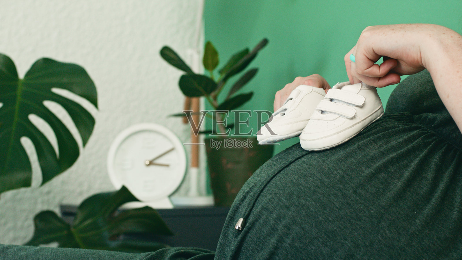 怀孕的母亲手握婴儿鞋，肚子上方被室内植物和时钟环绕照片摄影图片