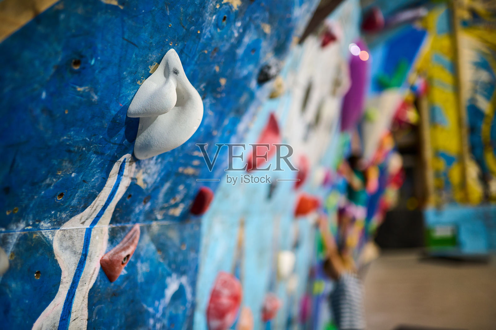 色彩丰富的室内攀岩墙上的白色攀爬把手特写，背景中模糊的年轻攀岩者。充满活力和能量的训练氛围。照片摄影图片