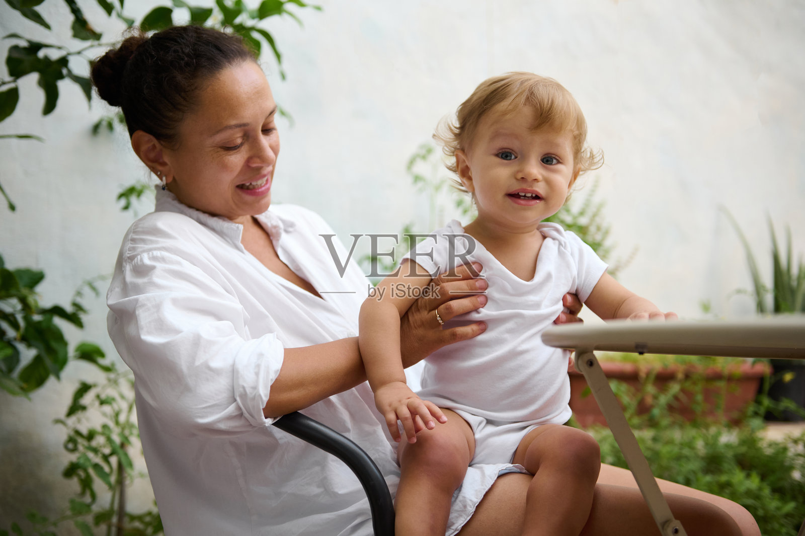 微笑的母亲在郁郁葱葱的花园中抱着她快乐的宝宝照片摄影图片