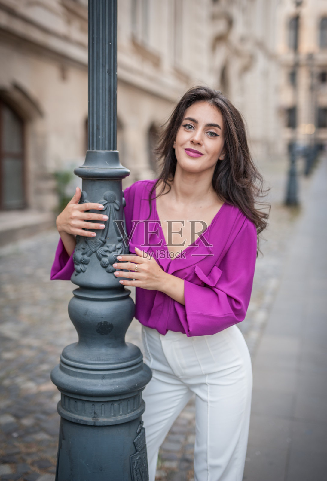 穿着紫色上衣和白色裤子的美丽年轻棕发女性在户外靠在柱子上摆姿势。照片摄影图片