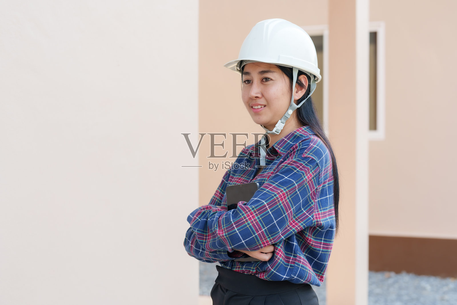 穿着格子衬衫的亚洲女性房屋检查员，戴着安全帽，双臂交叉，自信地站在住宅建筑工地外，展现出在住房项目中的专业性和权威性。照片摄影图片
