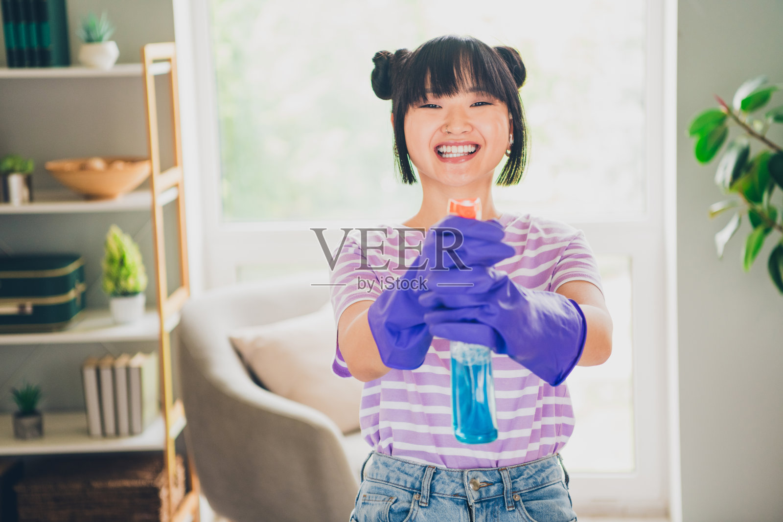 穿着紫色条纹T恤的积极心态女性在室内公寓房间里朝你喷洒喷雾。照片摄影图片