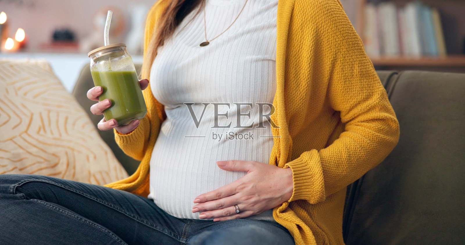 孕妇在沙发上手握绿色果昔，享受营养、消化健康和舒适。未来的母亲，肚子和手中拿着健康饮品，补充饮食维生素或增强免疫系统。照片摄影图片