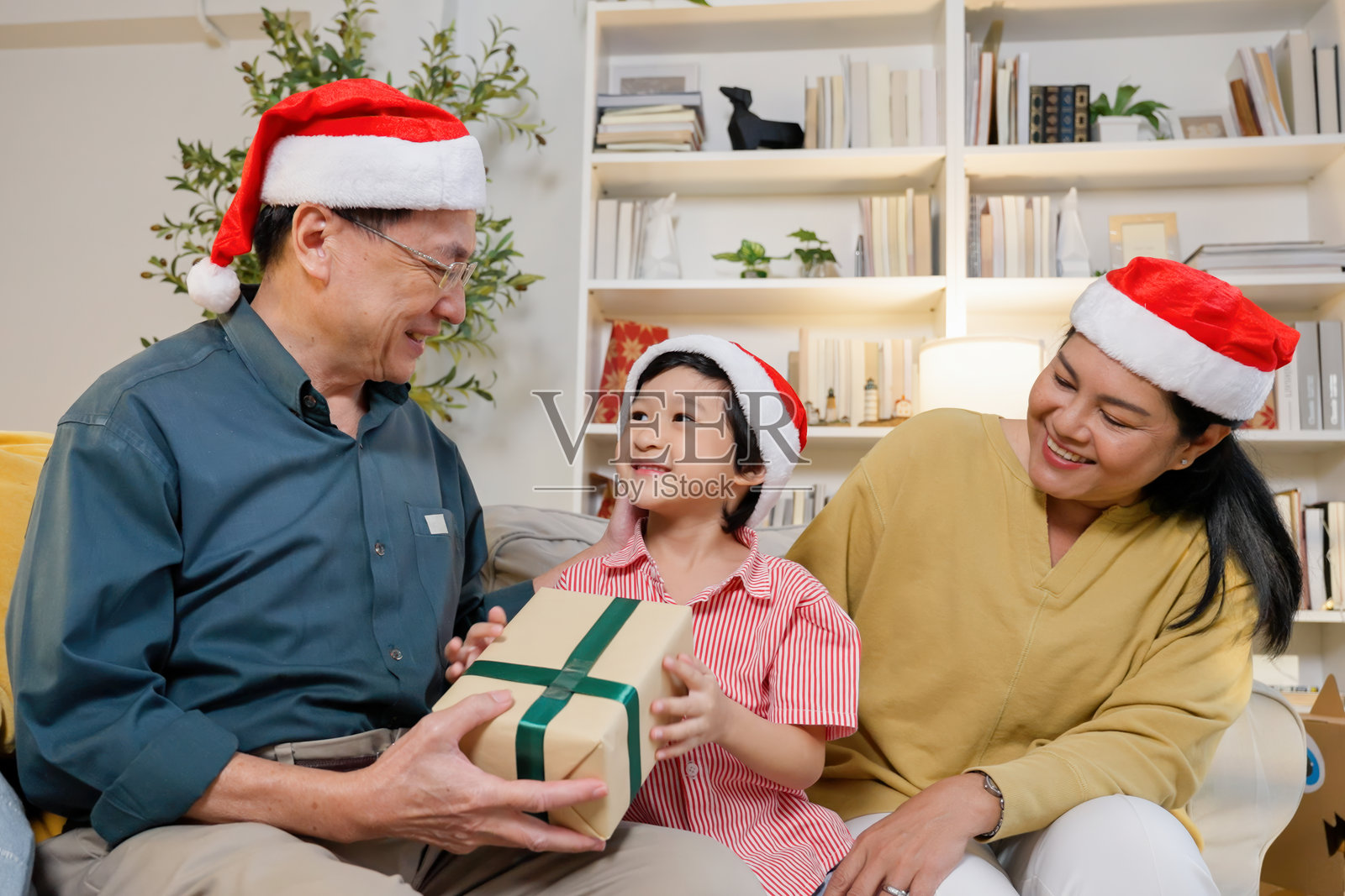亚洲祖父、祖母和孙子戴着圣诞帽在沙发上互赠圣诞礼物。温馨的节日氛围中展现出家庭的爱、快乐和联系，庆祝这个季节。照片摄影图片