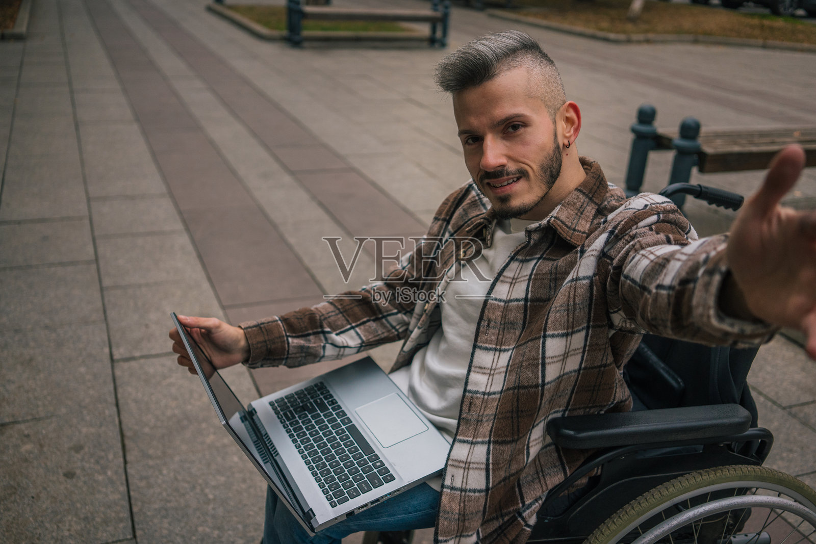 一位年轻快乐的轮椅男士在城市中悠闲地漫步，正用手机拍摄明亮的自拍照。照片摄影图片
