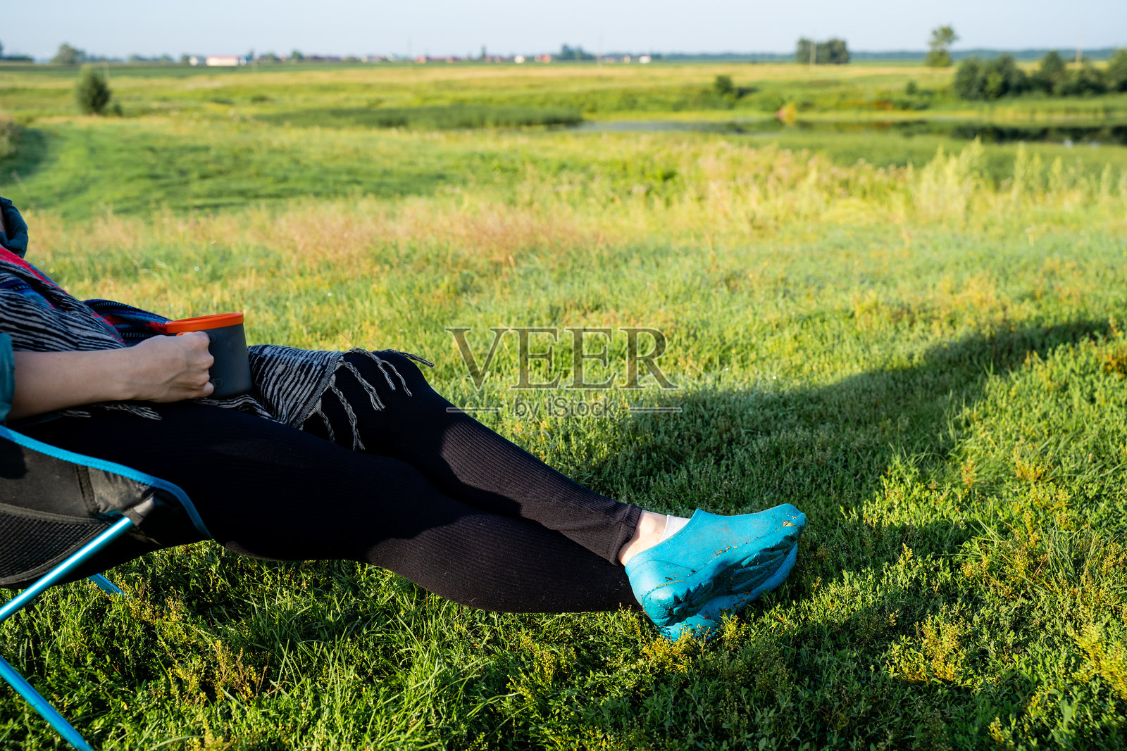 在宁静的风景中，一位女性舒适地坐在椅子上，双脚翘起，周围是一片生机勃勃的草地，天空湛蓝。照片摄影图片
