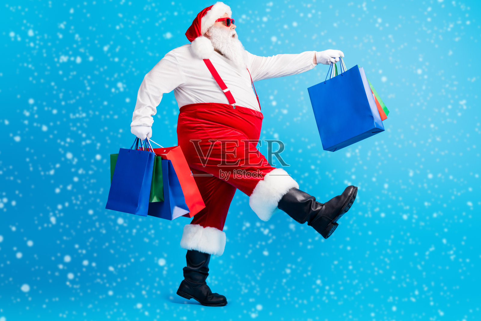 全身侧面照，疯狂的白灰色胡须胖圣诞老人正在购物，手里提着圣诞袋，穿着红色吊带工装，背景为蓝色。照片摄影图片