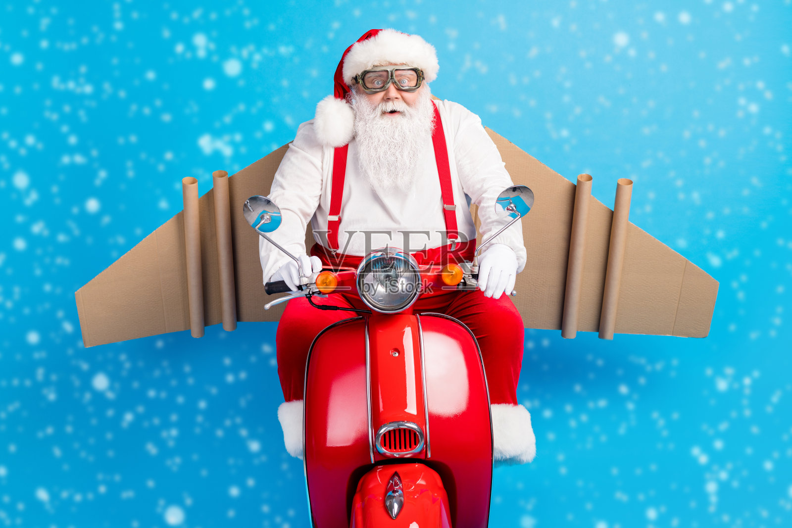 搞笑的疯狂圣诞老人骑着滑板车快速飞行，身穿现代时尚的吊带工装，戴着眼镜和护目镜，头戴帽子，背景为蓝色，身后有纸板制作的翅膀。照片摄影图片