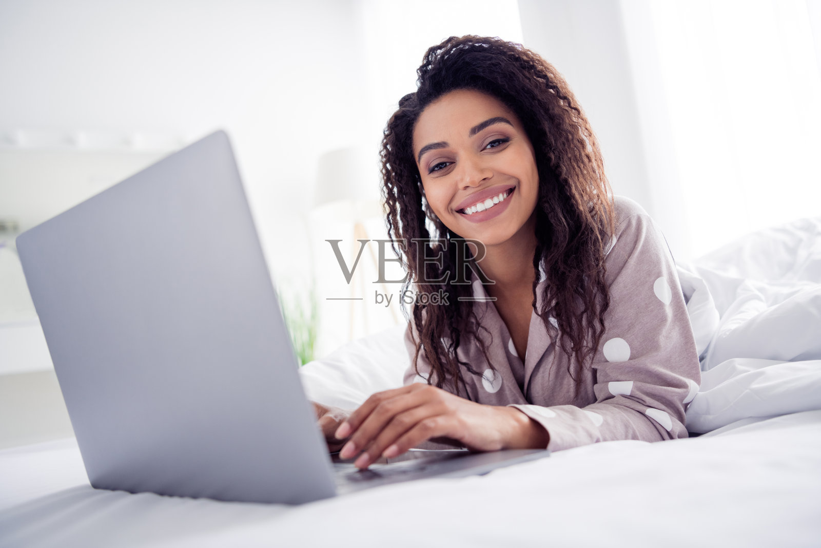 年轻女性穿着波点睡衣，微笑着在舒适的卧室里使用笔记本电脑。照片摄影图片