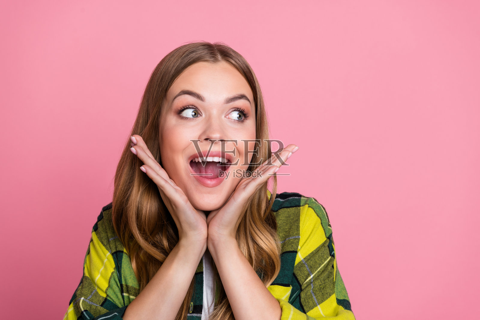 穿着时尚黄色格子衣服的迷人年轻女性，兴奋地看着空旷的空间，背景为粉色。照片摄影图片