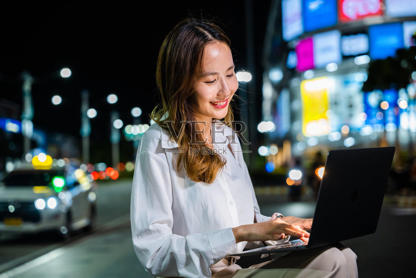 当城市的灯光照亮夜空时，一位女性正专心致志地在她的笔记本电脑上工作。照片摄影图片
