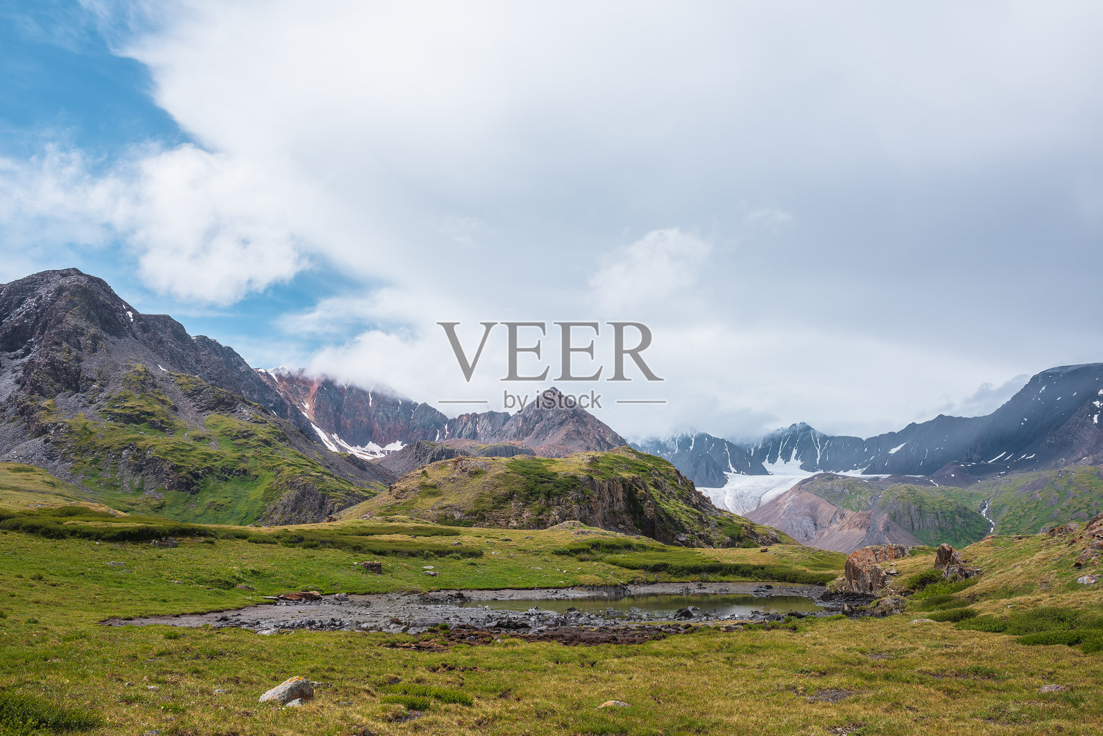 绿丘和岩石间的阿尔卑斯湖，背景是尖锐的山峰和高耸的山脊，笼罩在低云之中。雨雾弥漫的天空下，壮观的冰川舌和大山脉的轮廓若隐若现。照片摄影图片