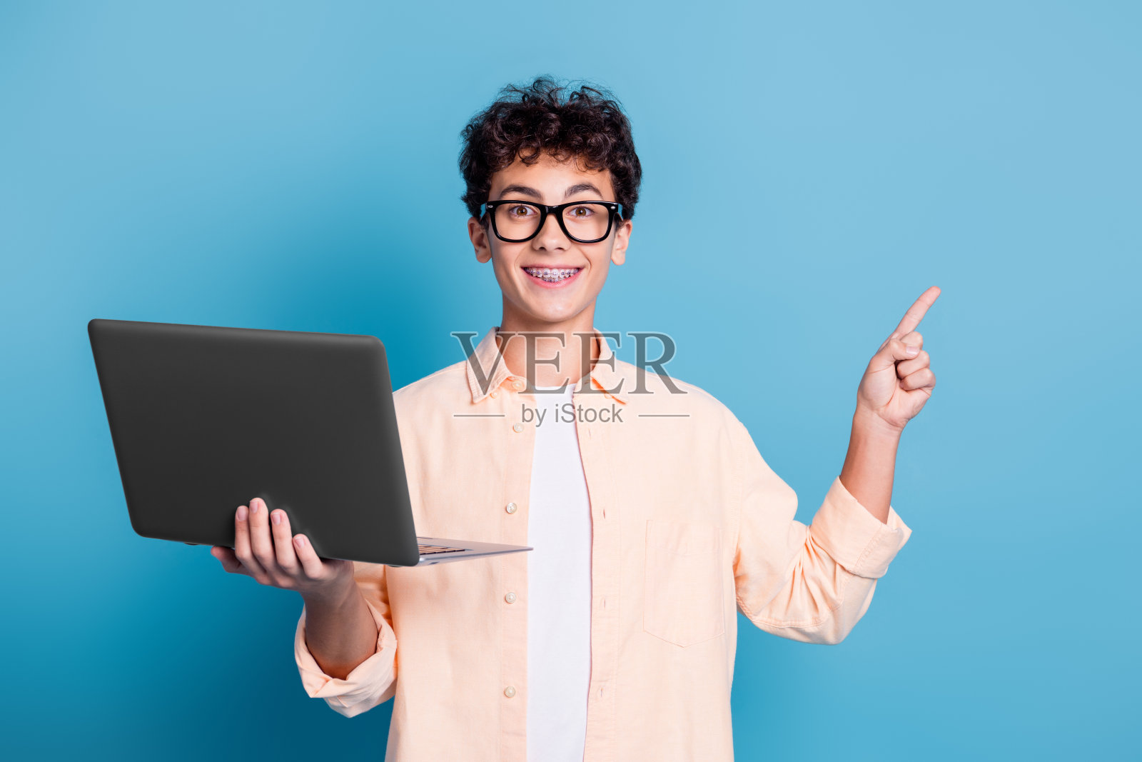 微笑的年轻男子戴着牙套，手持笔记本电脑并指着，穿着休闲衬衫，站在蓝色背景前。照片摄影图片