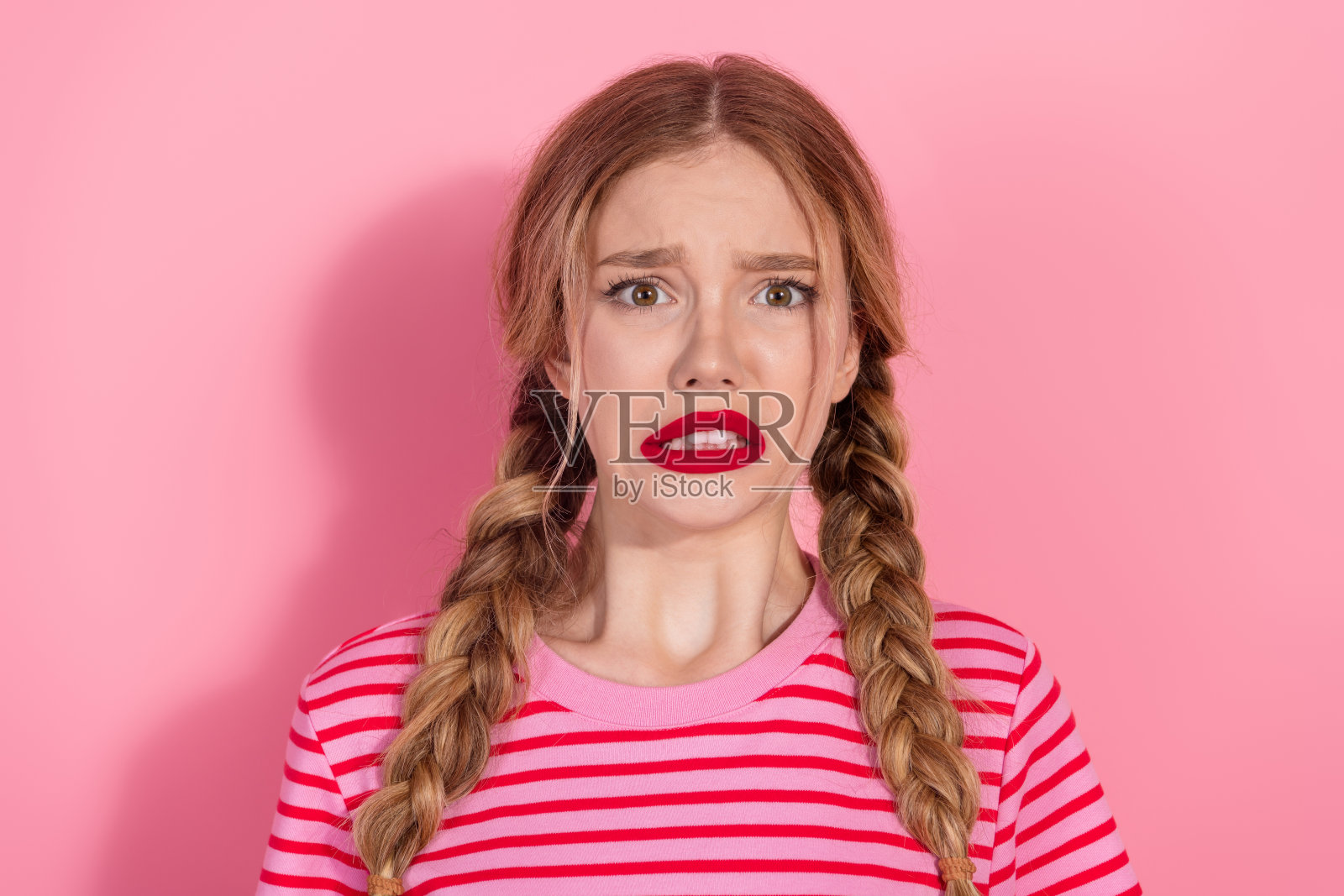 困惑的年轻女性，扎着辫子，涂着大胆的红唇，穿着时尚的夏季服装，站在粉色背景前。照片摄影图片