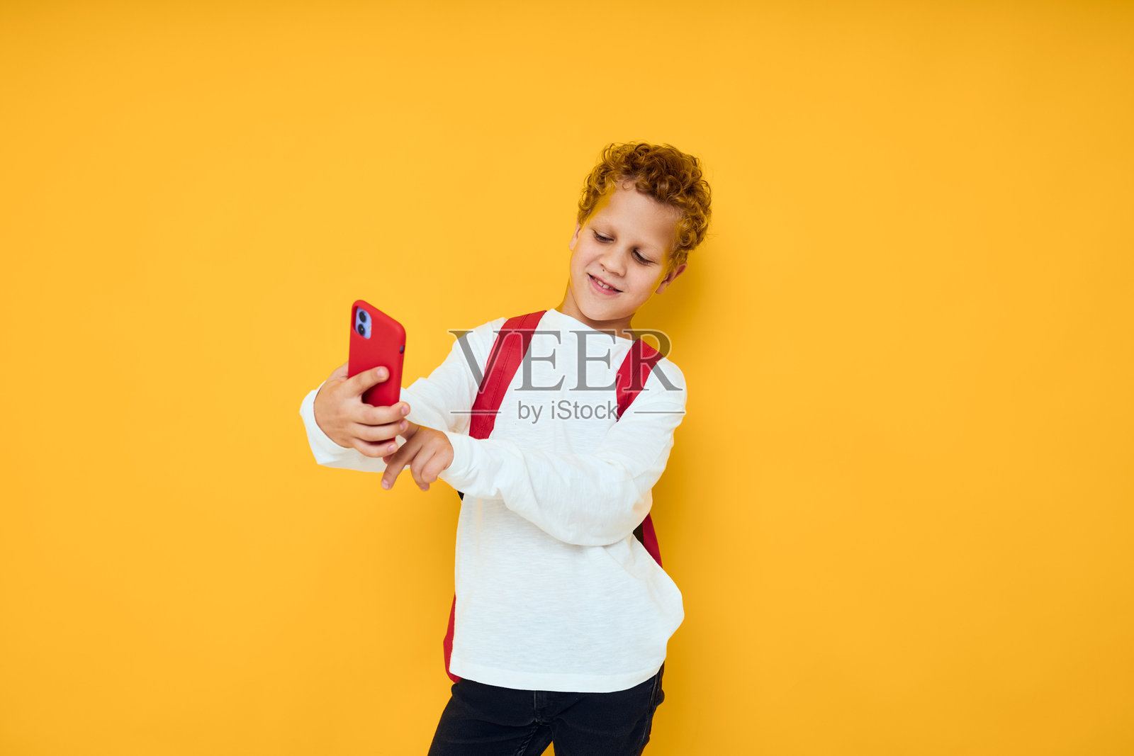 穿着白色卫衣的搞笑男青少年，手持手机，背着红色背包，背景为纯色。照片摄影图片