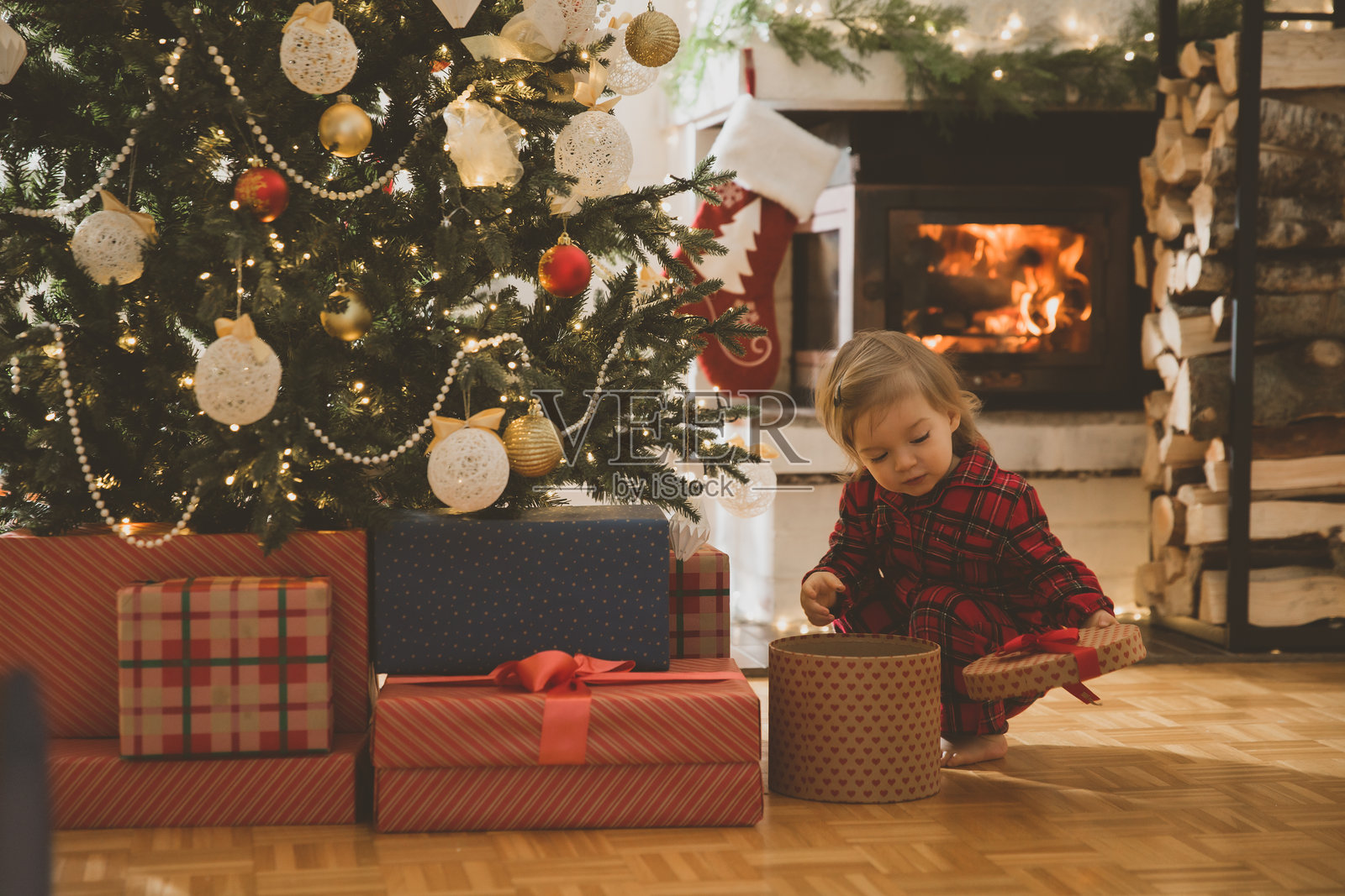 在家中传统庆祝圣诞节。可爱的宝宝穿着红色格子睡衣在装饰好的圣诞树下拆礼物。美丽舒适的房子里有壁炉。低调的电影灯光。照片摄影图片
