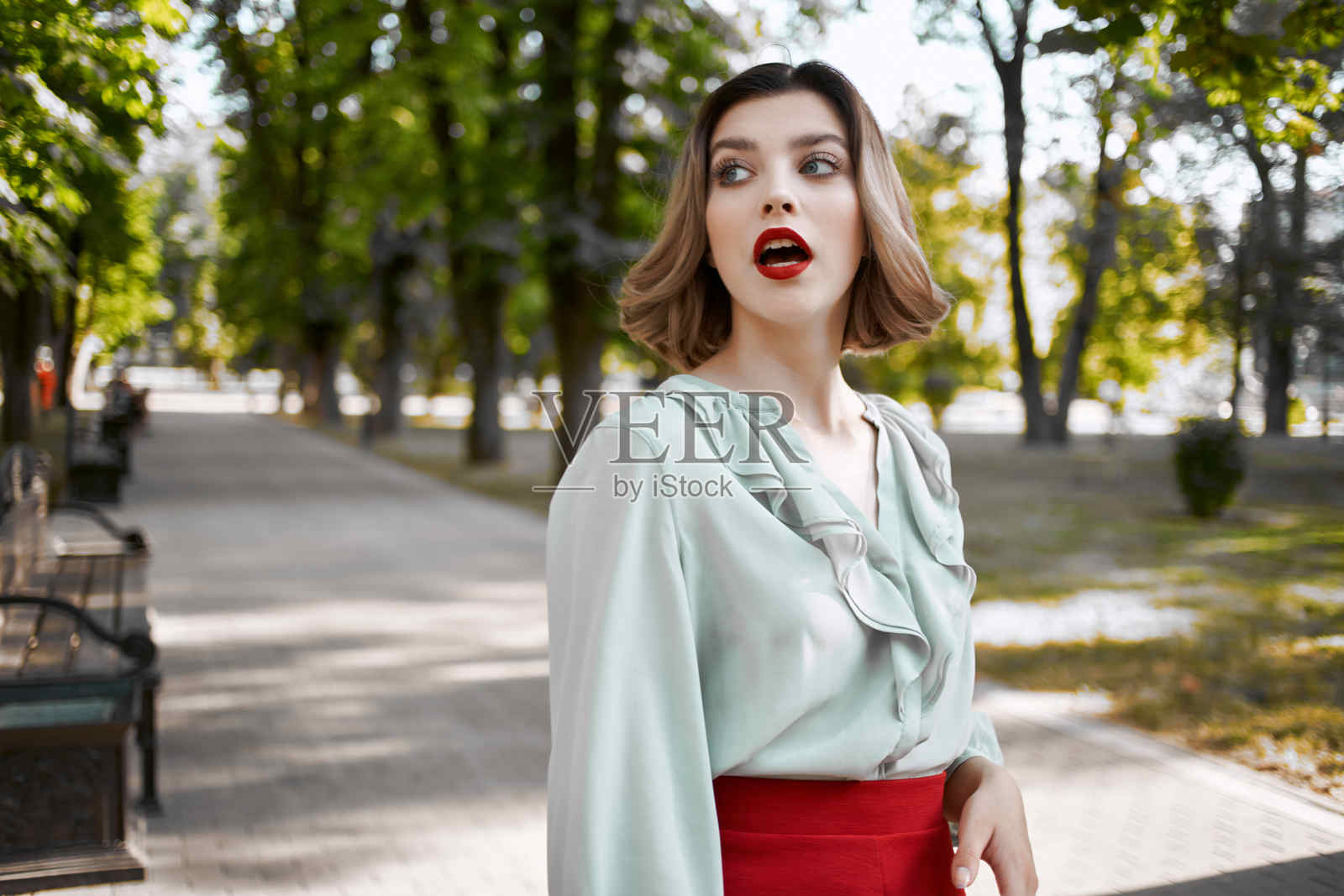 穿着红色裙子的漂亮金发女孩在公园的绿树下。照片摄影图片