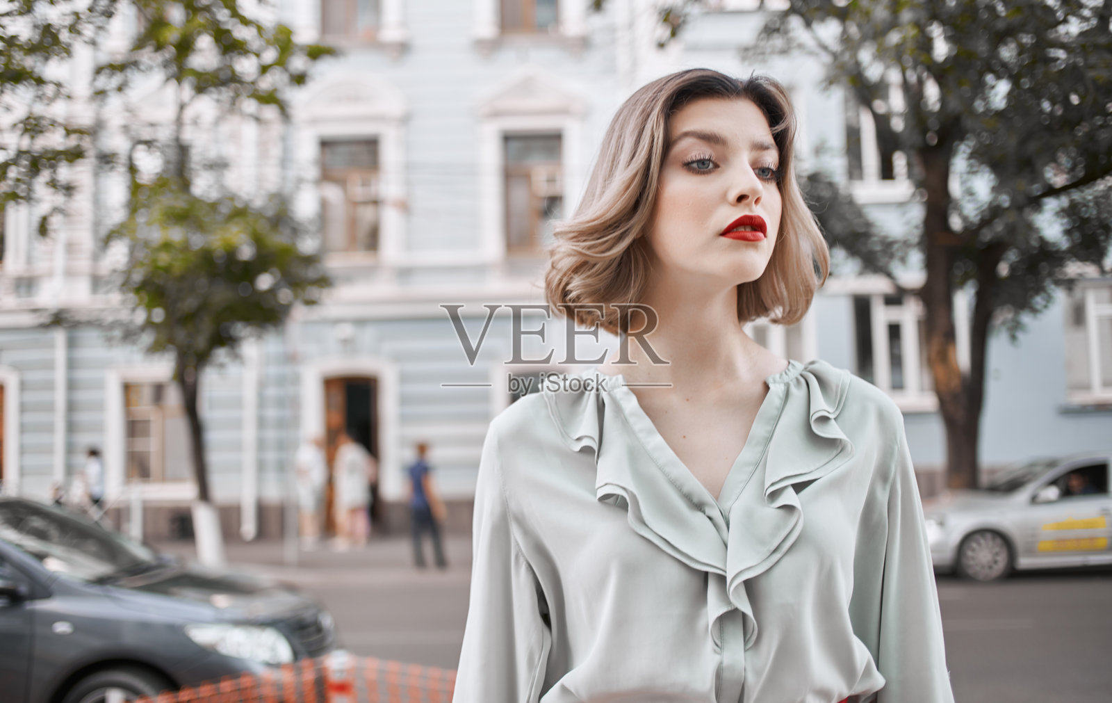 穿着时尚衬衫、红唇的女性在建筑物和绿色树木旁，春天的自然景色。照片摄影图片