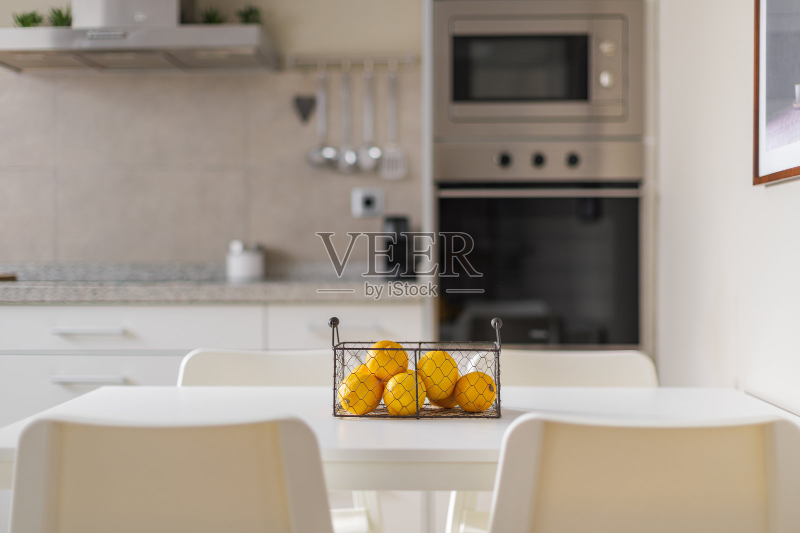 金属篮子里装着成熟的黄色柠檬，放在白色餐桌上。背景模糊的厨房家具和墙上刷过的耐用金属电子设备。照片摄影图片