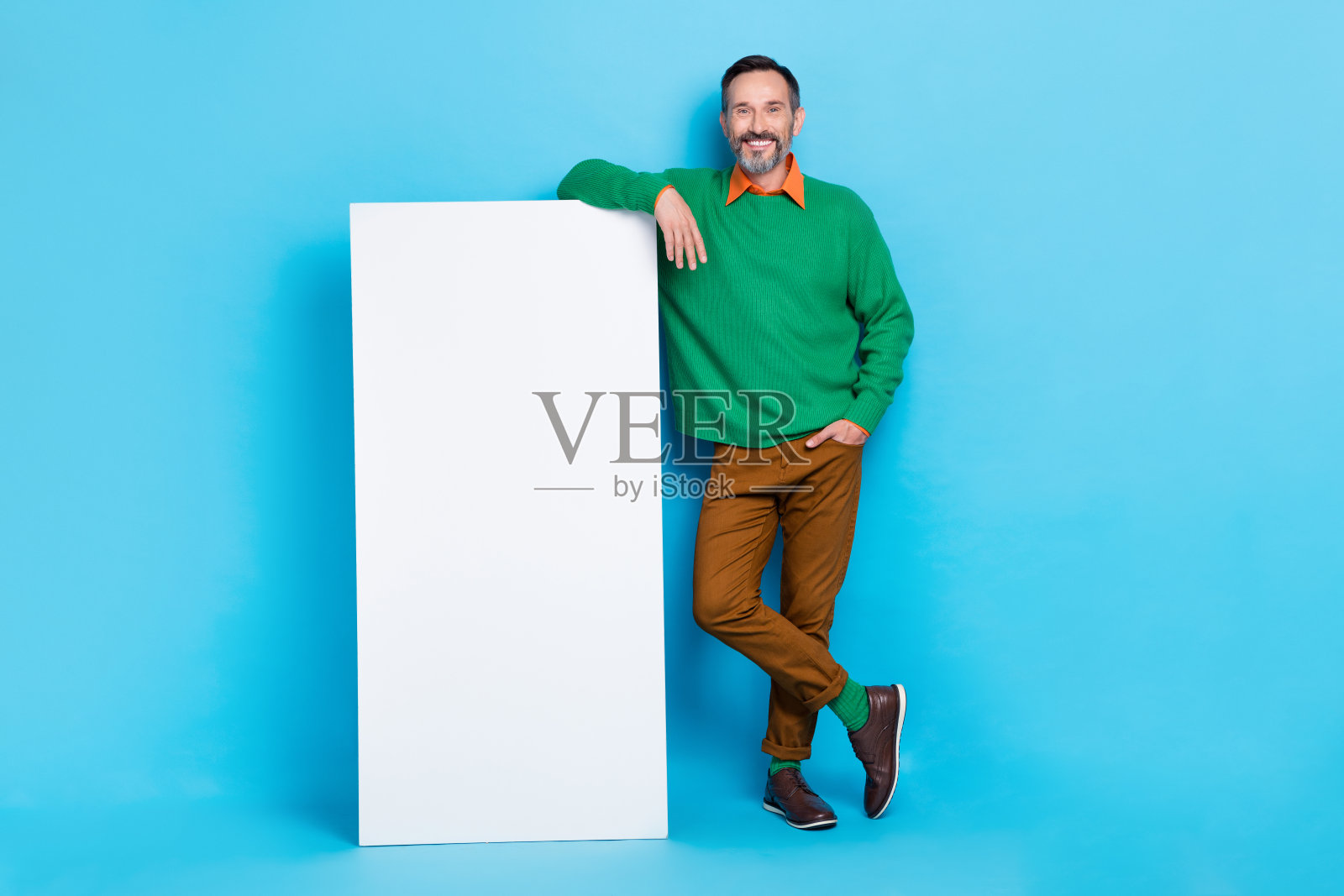 成熟帅气的男人全身照，靠在垂直的白色海报横幅上，穿着时尚的绿色衣服，背景为蓝色。照片摄影图片