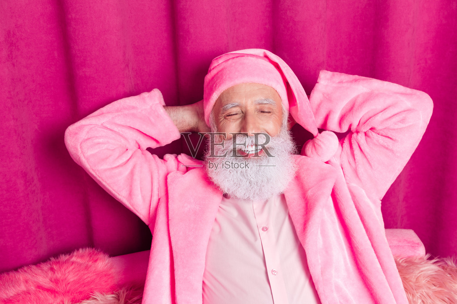 老年圣诞老人穿着浴袍，戴着帽子，躺在粉色沙发上休息，房间窗帘的室内环境。照片摄影图片
