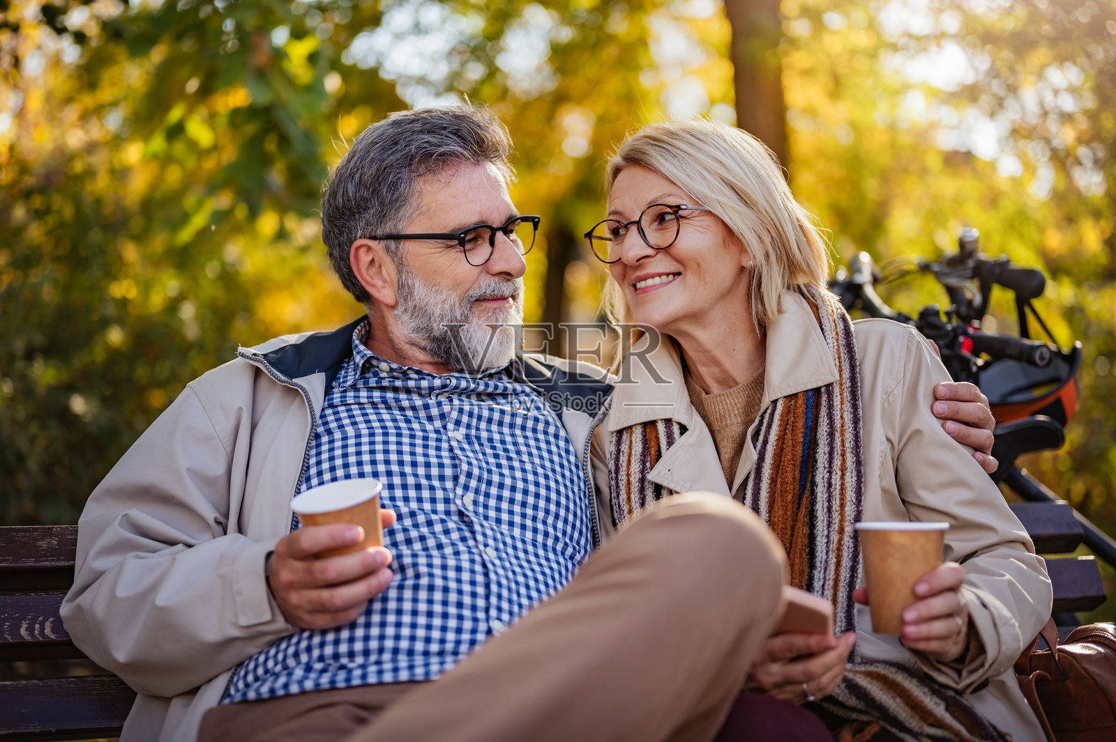 快乐的老年夫妇坐在公园长椅上，互相拥抱，一起享受阳光明媚的秋日咖啡，背景中有自行车。照片摄影图片