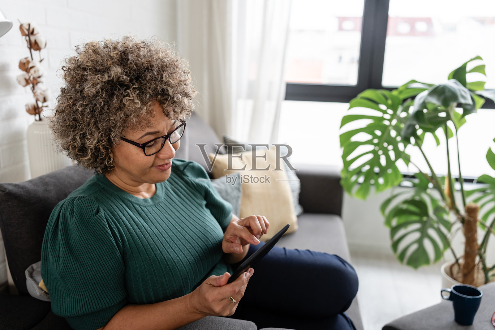 一位中年女性，卷发，戴着眼镜，穿着绿色毛衣，坐在大窗户旁，微笑着在电子书阅读器上阅读，享受着在家放松的时光。照片摄影图片