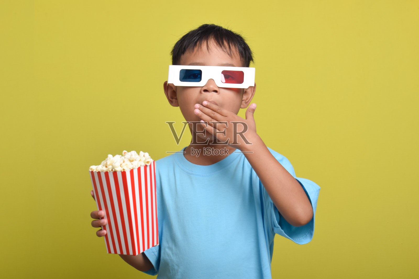 震惊的亚洲男孩，双手捂住嘴，戴着3D眼镜，手里拿着爆米花桶照片摄影图片