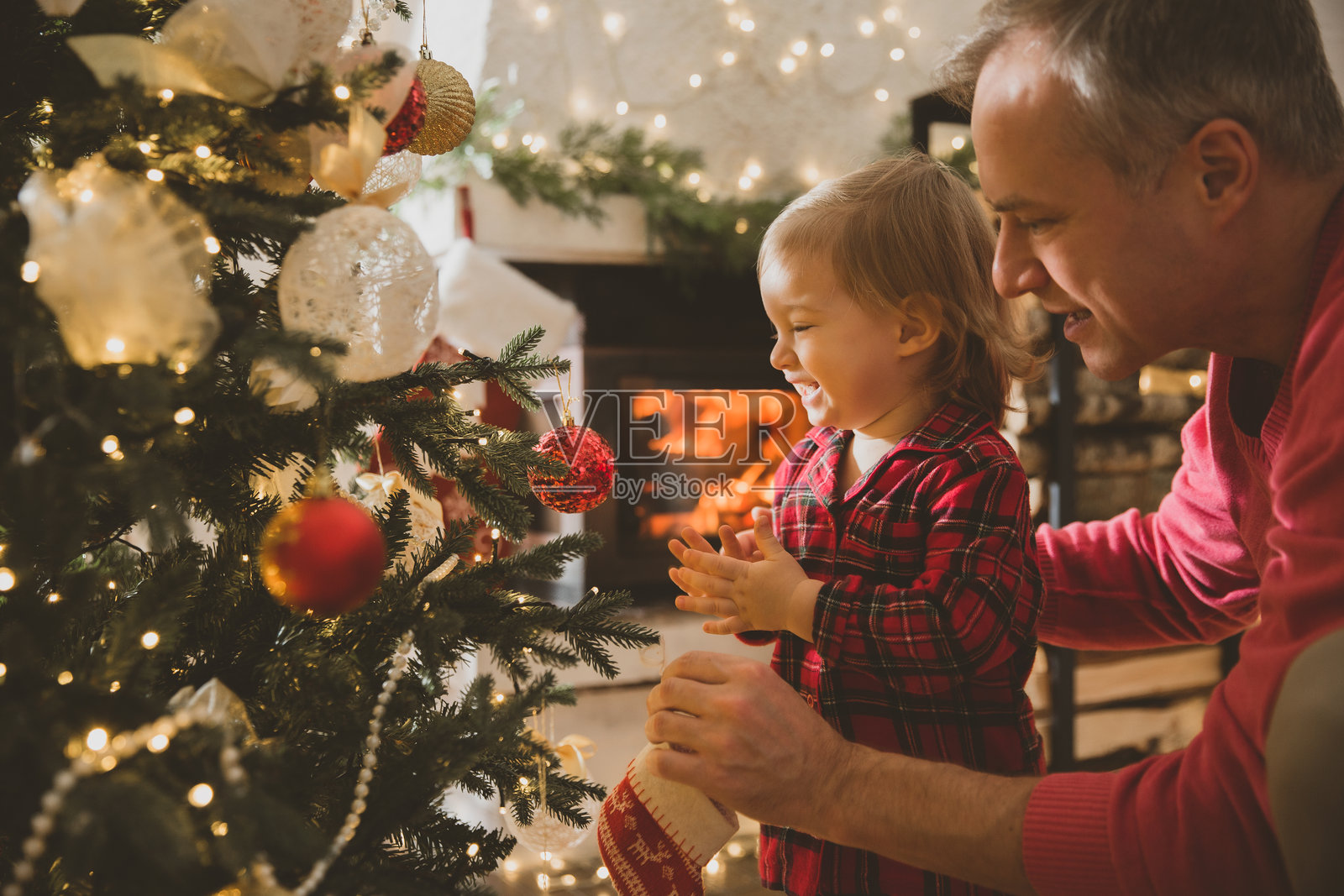 父亲和他兴奋的幼儿在家庆祝圣诞节。家人们一起快乐地装饰圣诞树。可爱的宝宝穿着红色格子睡衣。美丽的房子里有壁炉，地上摆满了包好的礼物。照片摄影图片