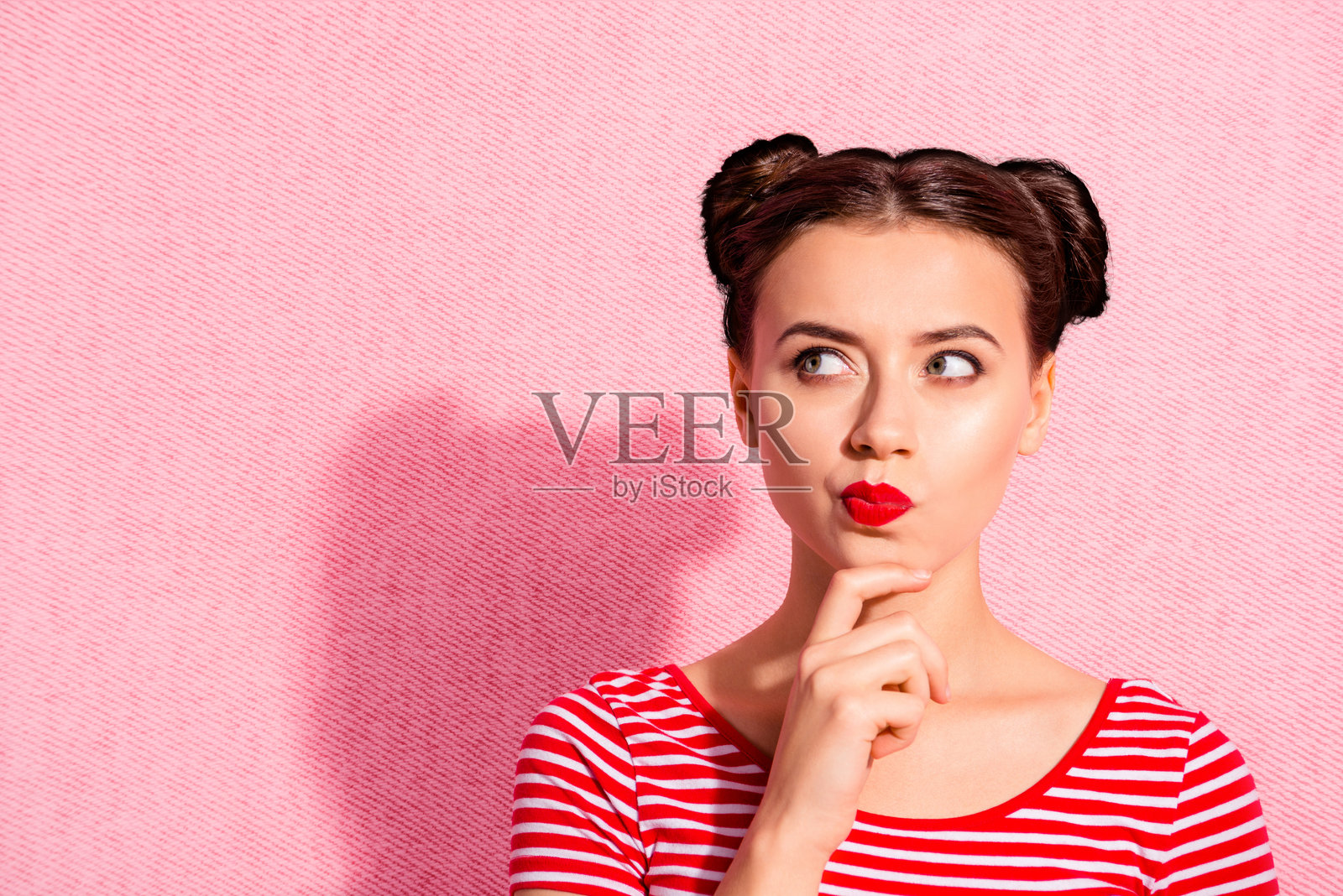 特写照片：美丽的女士，两个发髻，涂抹唇膏，手托下巴，仰望空旷的空间，沉思 contemplative，穿着休闲的红白条纹T恤，背景为粉色。照片摄影图片