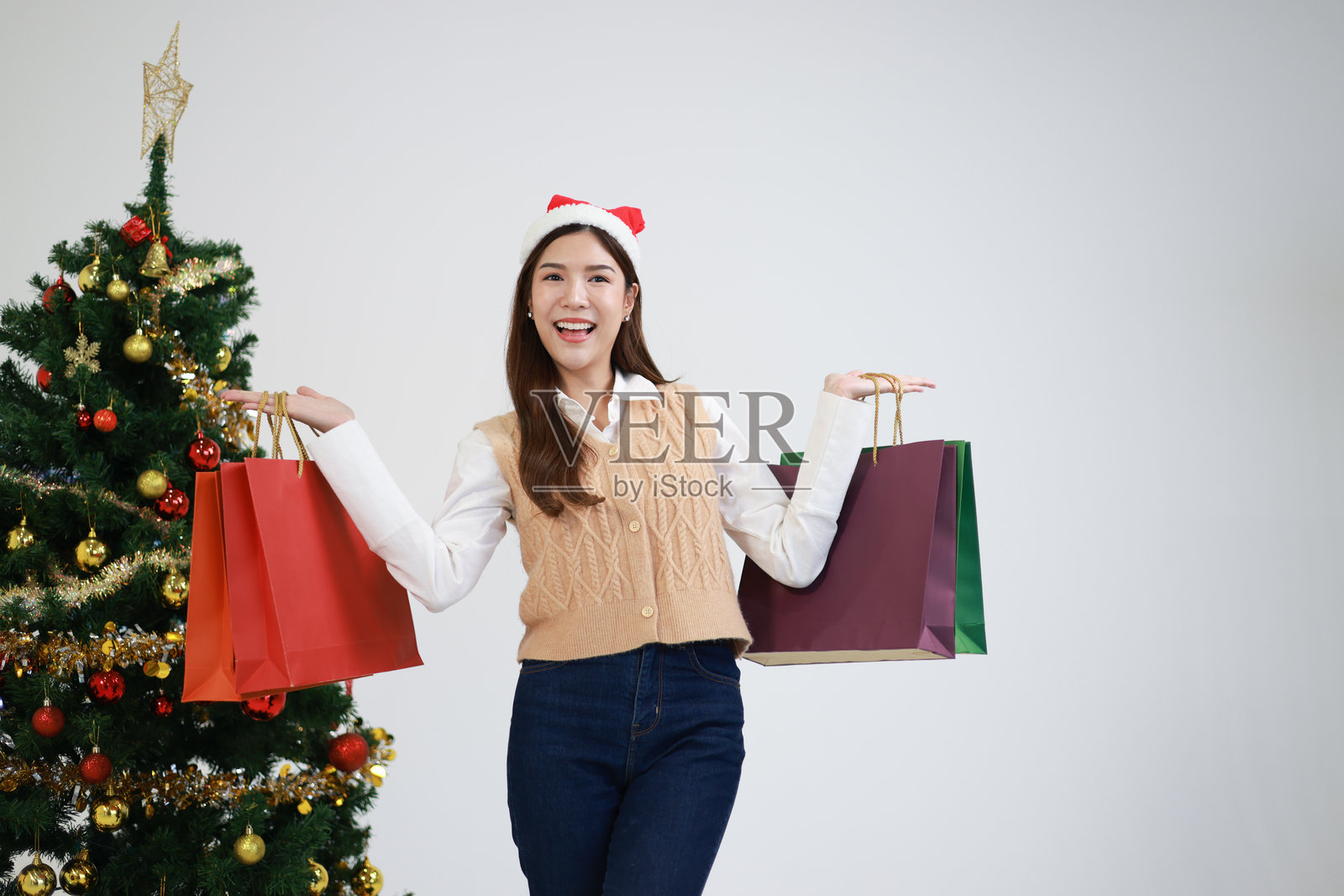 微笑的女性穿着圣诞老人服装，手里提着圣诞购物袋和礼物，站在装饰好的圣诞树旁。照片摄影图片