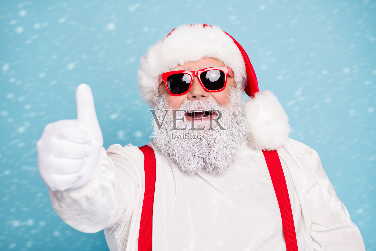 特写照片，搞笑的胖胖圣诞老人戴着现代的时尚眼镜，竖起大拇指推荐冬季促销，穿着红色吊带工装，背景为蓝色。照片摄影图片
