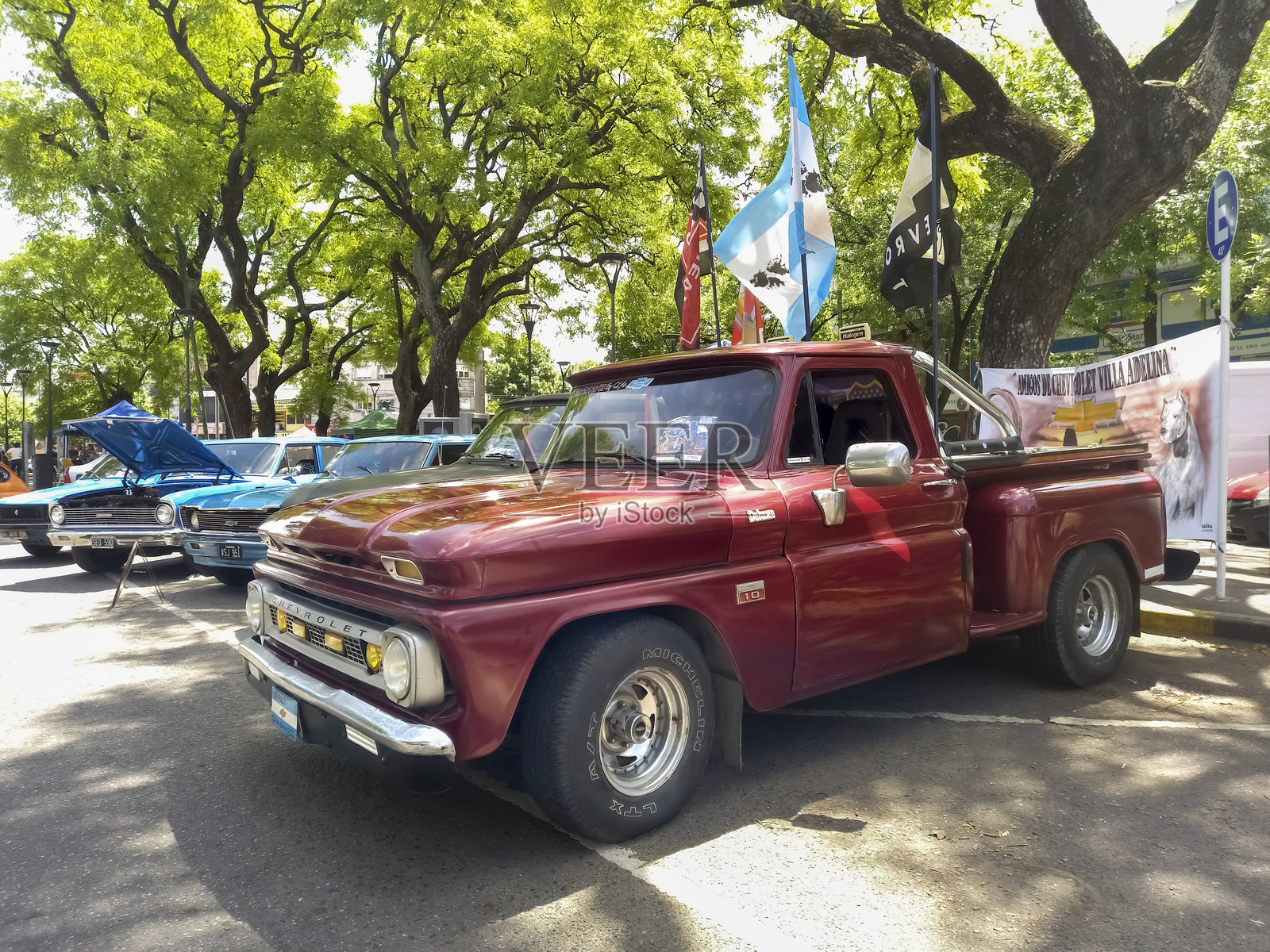 1960年代中期的老红色雪佛兰C10皮卡车停在城市街道的树下，参加经典汽车展。照片摄影图片