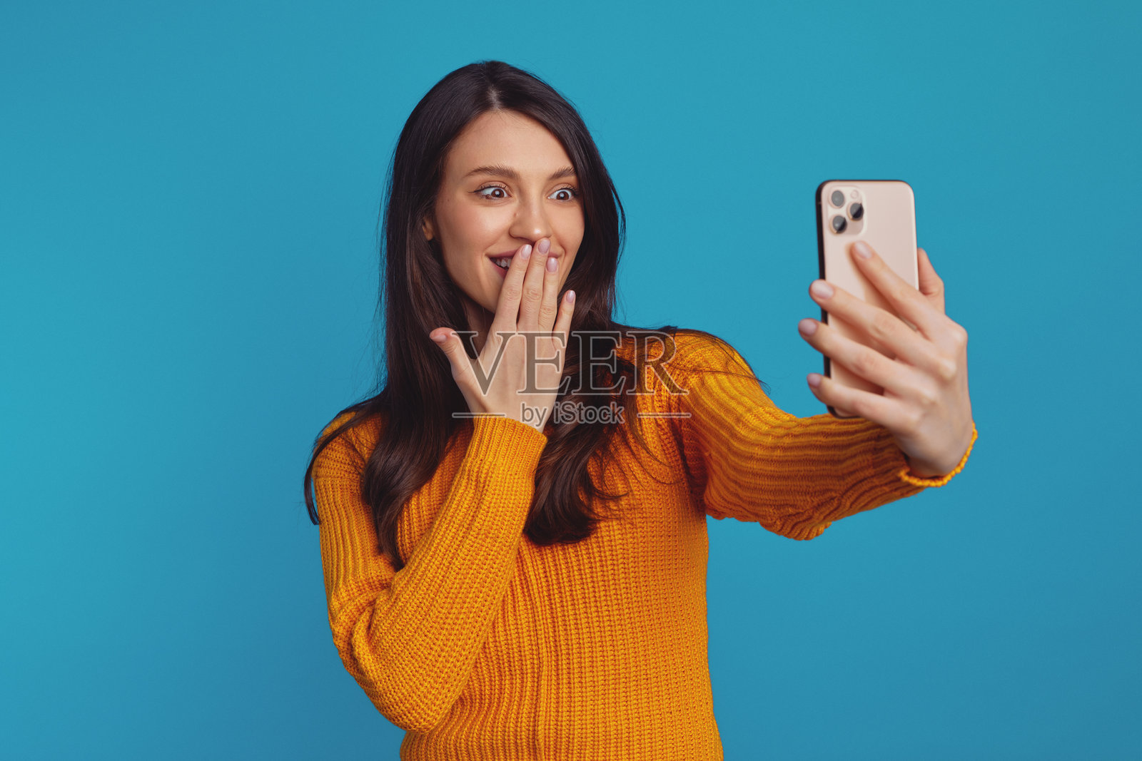 穿着橙色毛衣的可爱女孩在蓝色背景前用智能手机自拍照片摄影图片