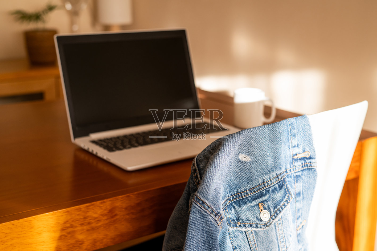 阳光明媚的极简家居工作空间，配有笔记本电脑、木质家具和休闲风格。舒适的家庭办公室角落，桌上有笔记本电脑，休闲牛仔夹克搭在椅子上。照片摄影图片