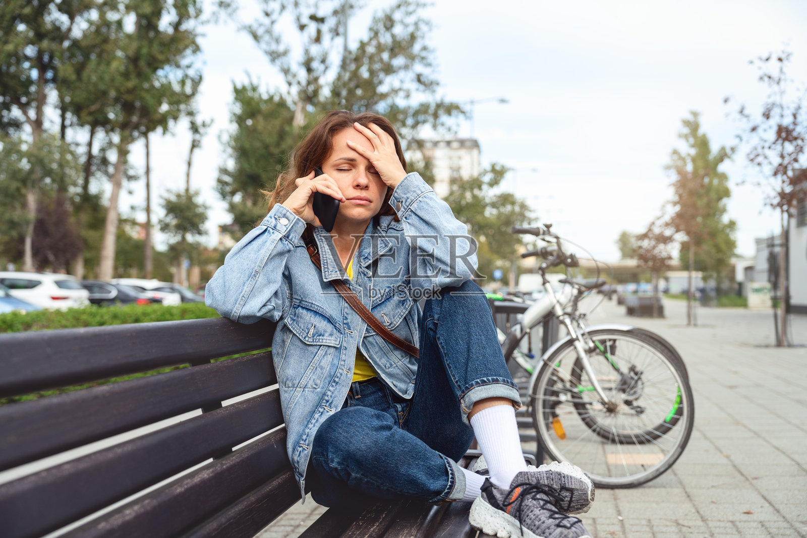 城市女性坐在长椅上，手扶额头，面露痛苦神情，正在手机上听取坏消息。照片摄影图片