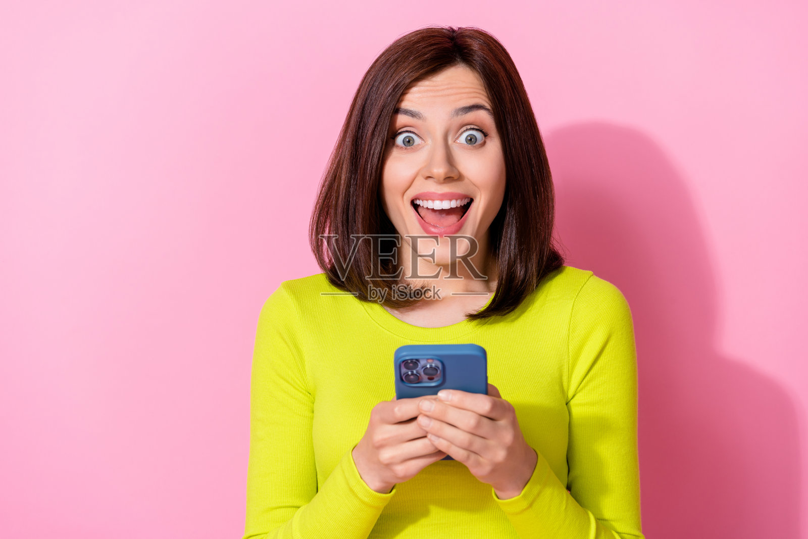 兴奋的漂亮女士穿着黄色上衣，张嘴聊天，手持现代电子设备，背景为粉色。照片摄影图片
