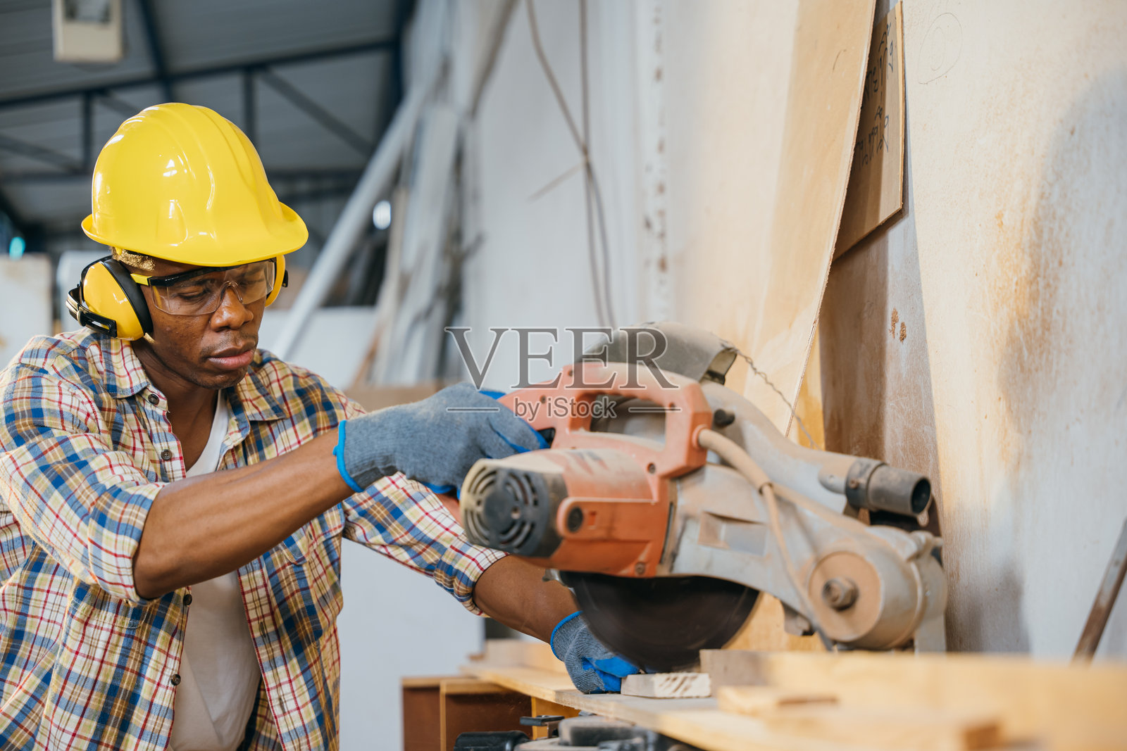 戴着黄色安全帽的建筑工人在使用切割锯切割木材，旁边有吸尘器在收集木屑。木工、精密切割和建筑工地照片摄影图片