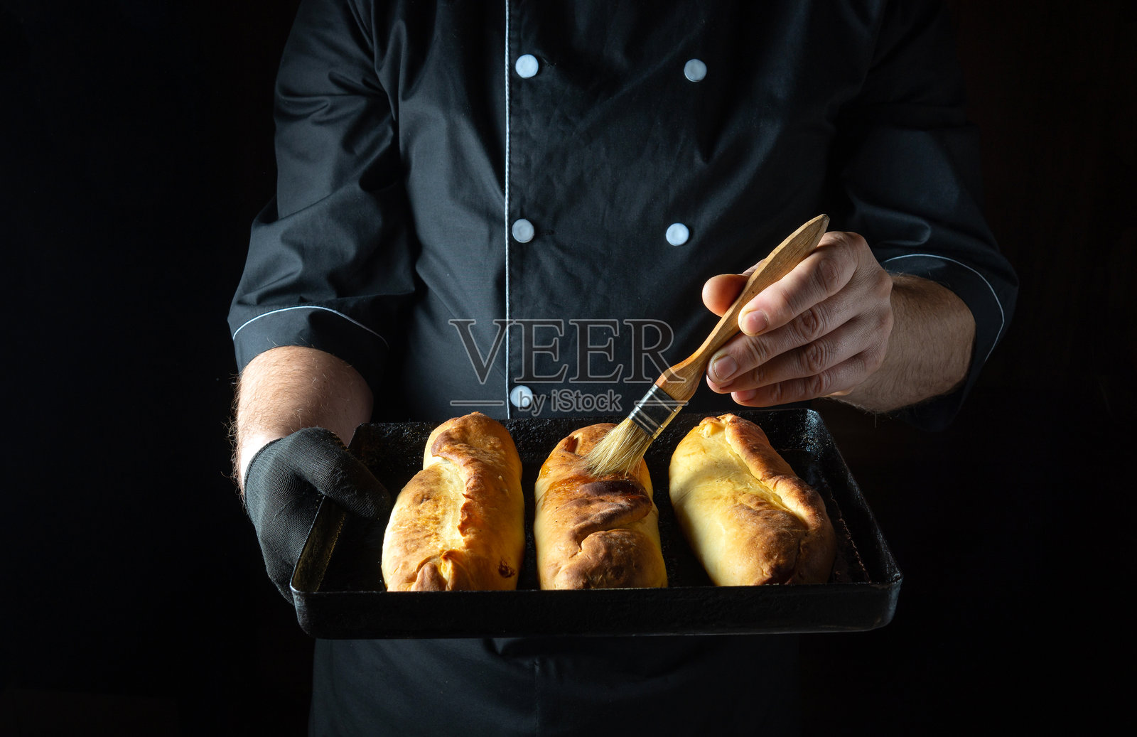 一位熟练的面包师小心翼翼地在三只金黄色的面包卷上刷上光泽，展现出它们酥脆的质地和诱人的香气。温暖的氛围散发着舒适感和烹饪热情。照片摄影图片