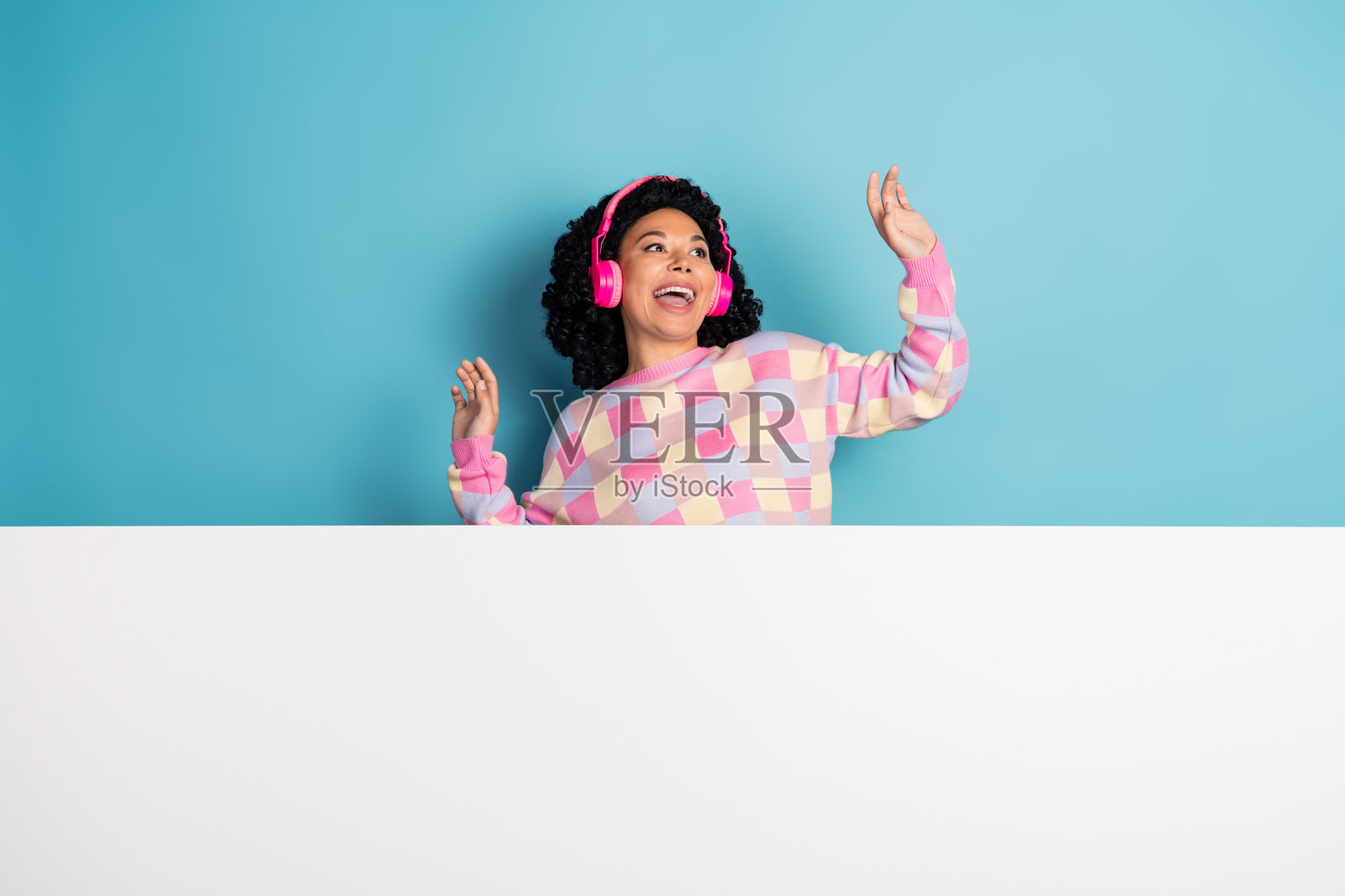 漂亮年轻女孩戴着耳机在蓝色背景下跳舞，穿着时尚的粉色格子服装，白色空旷空间中的照片肖像。照片摄影图片