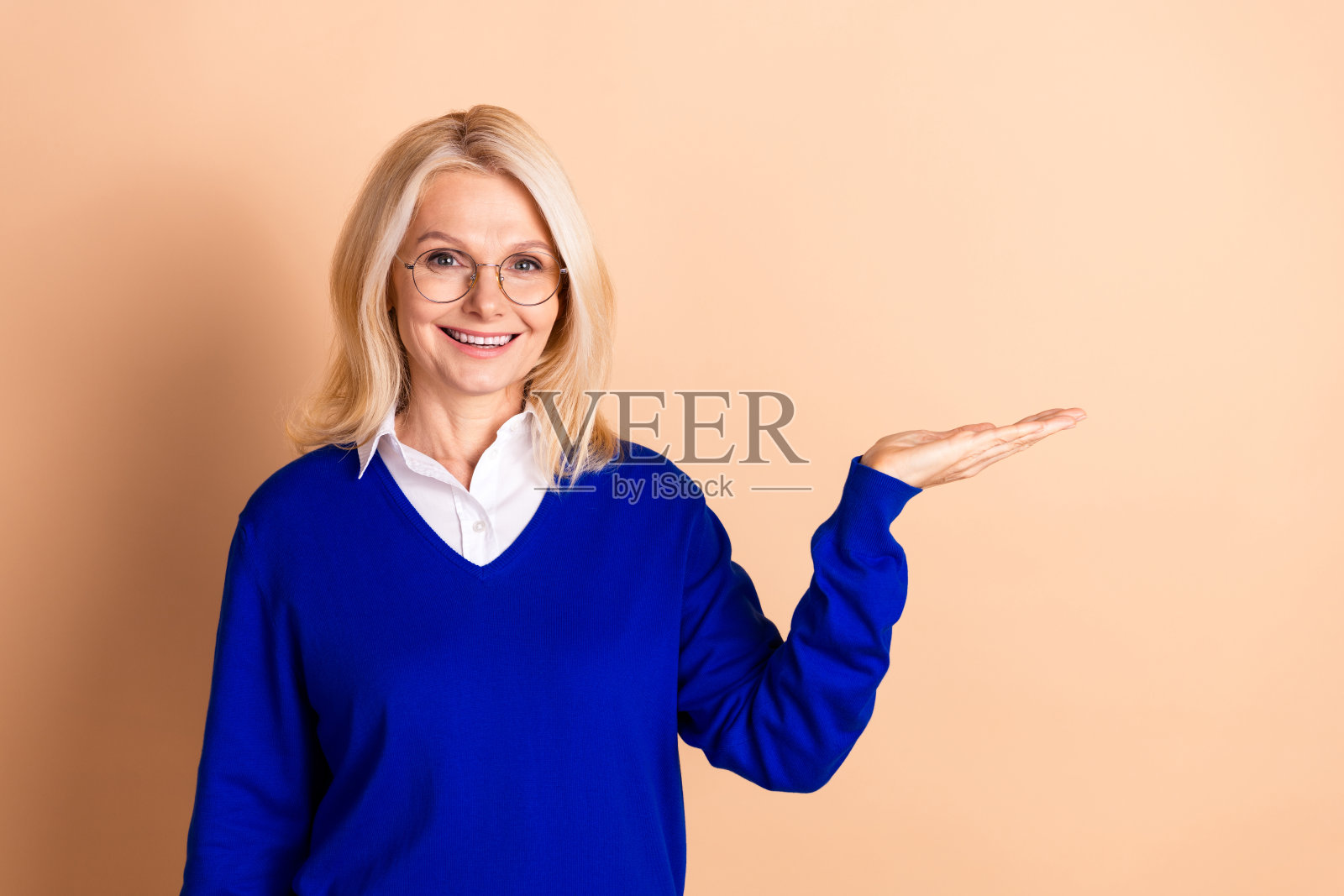 自信的成熟的女商人戴着眼镜，穿着蓝色毛衣在米色背景前展示。照片摄影图片