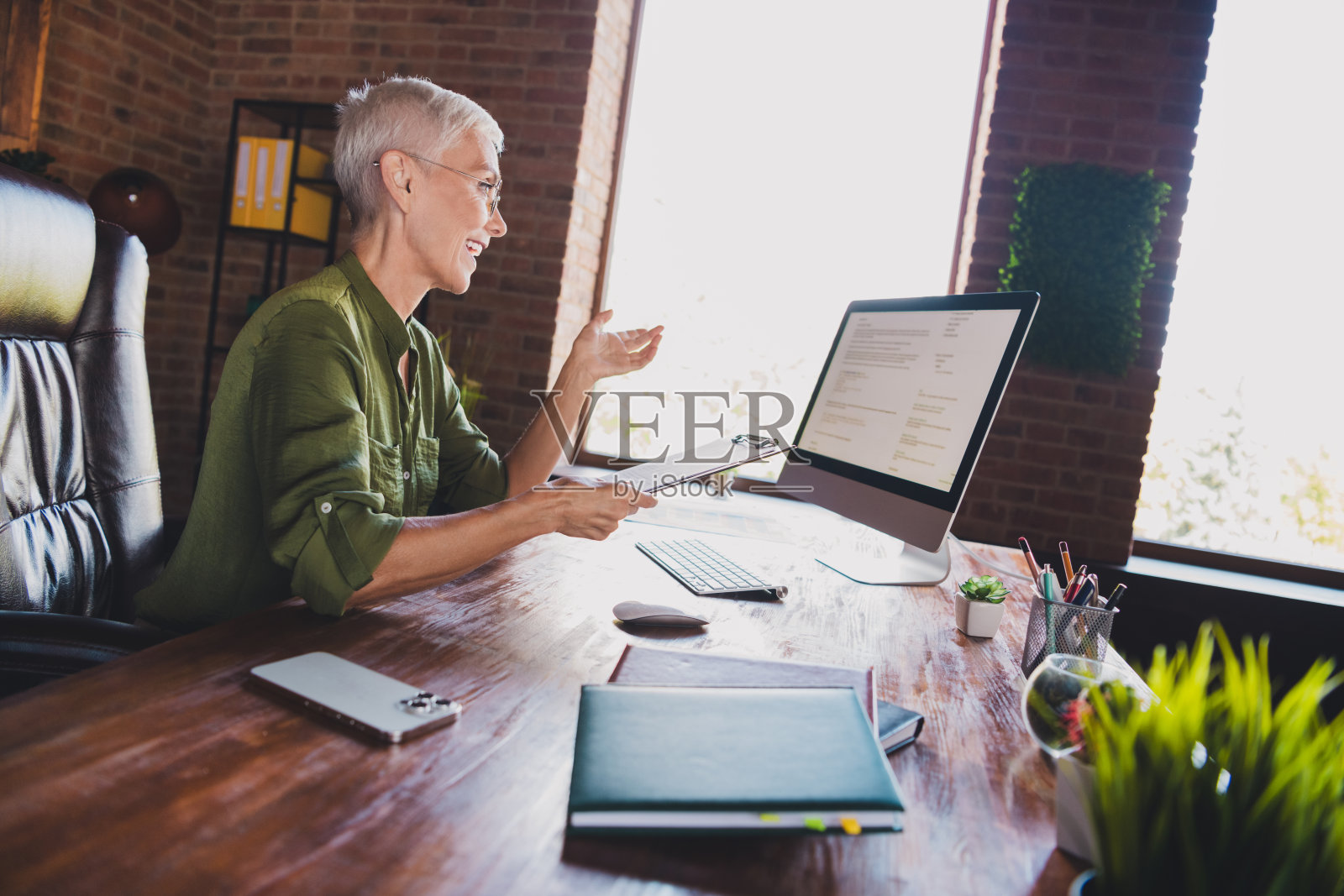 成功的年长女商人手持夹板，电脑显示屏前，穿着衬衫，现代阁楼室内办公室环境。照片摄影图片